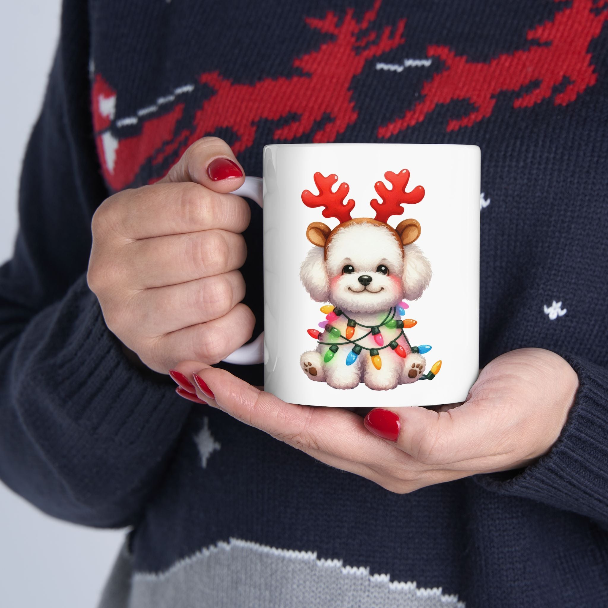 A person in a festive sweater holds a white mug with a Bichon Frise wearing reindeer antlers.