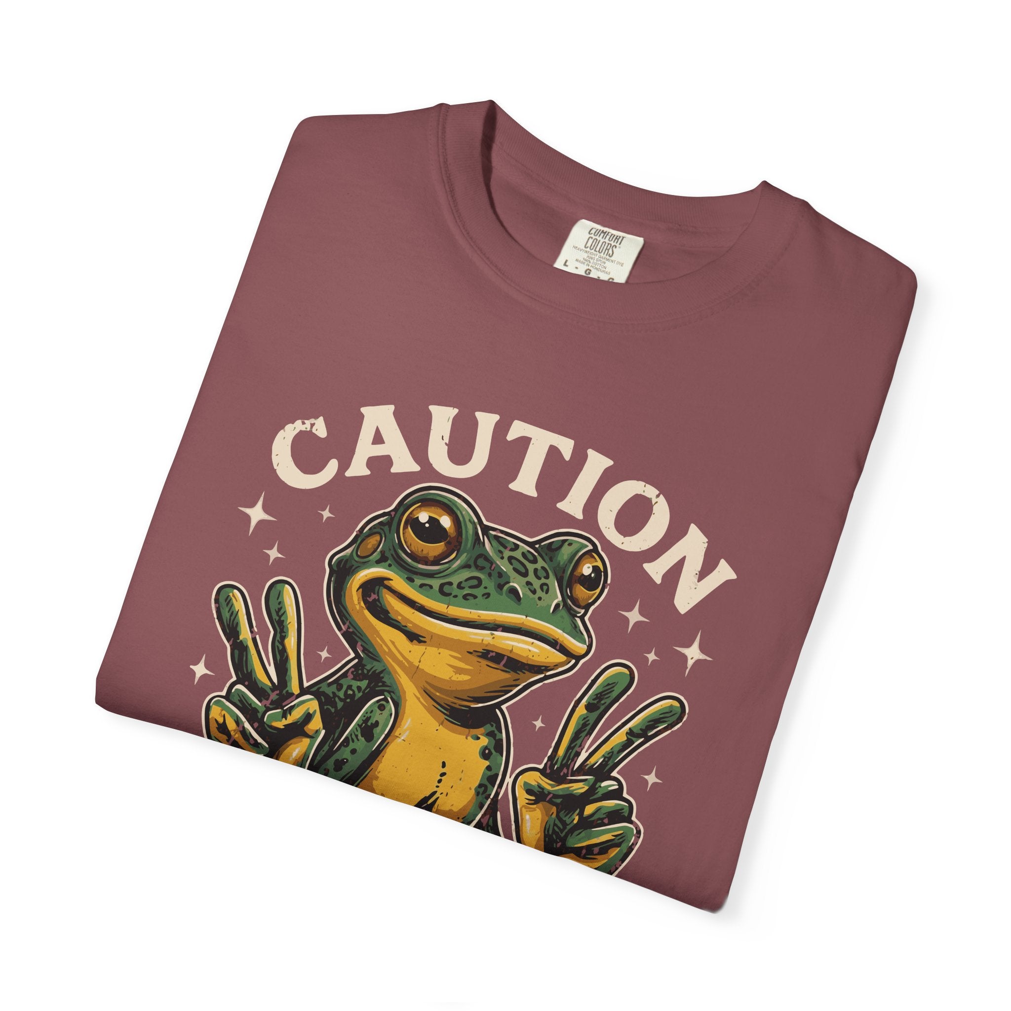 A folded maroon t-shirt featuring a graphic of a frog making peace signs with the word CAUTION.