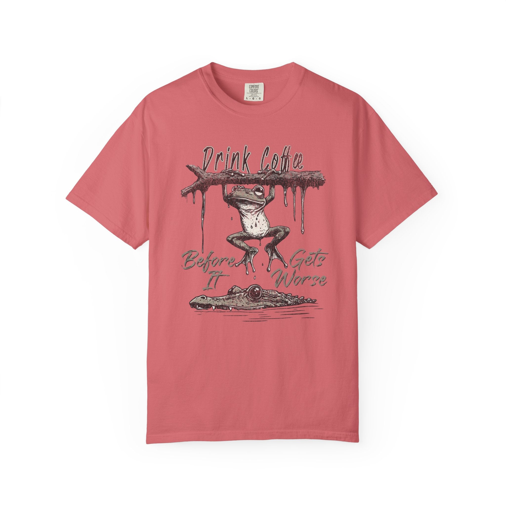 A coral-colored unisex t-shirt featuring a graphic of a frog hanging from a branch above a gator.
