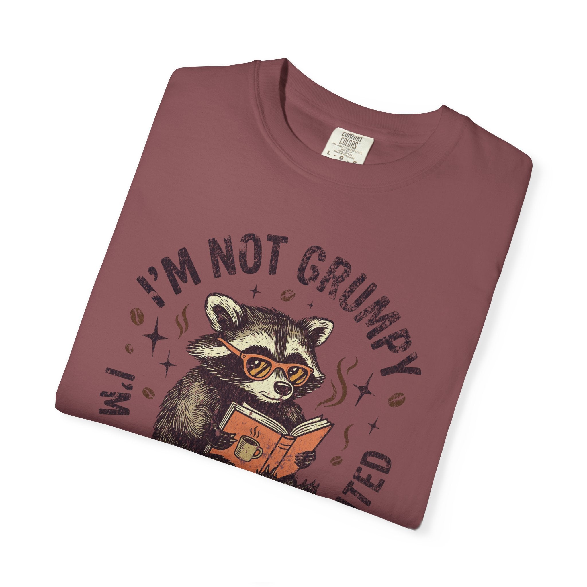 A maroon unisex t-shirt featuring a graphic of a raccoon wearing glasses and reading a book.