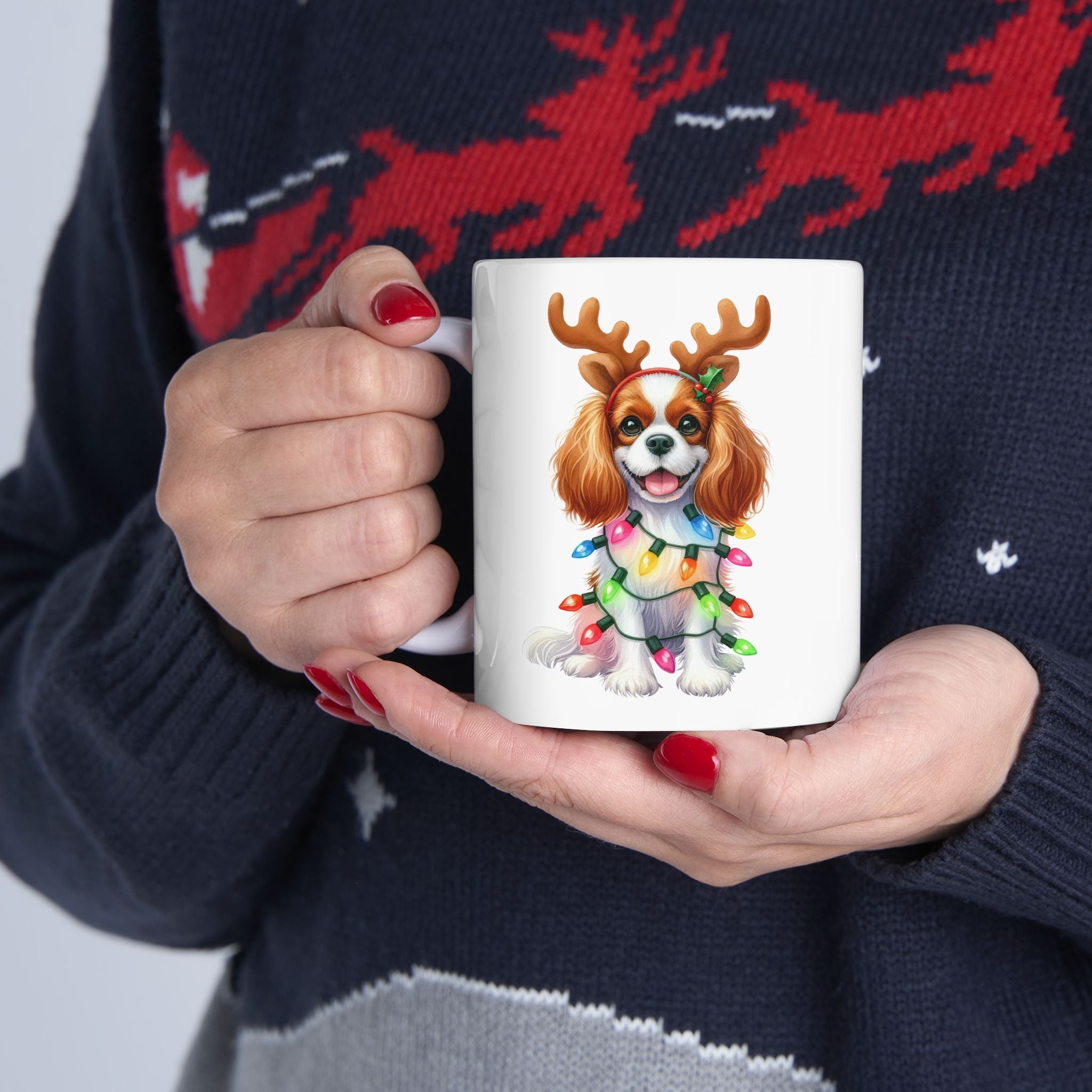 A person in a reindeer sweater holds a white mug with a Cavalier King Charles Spaniel.