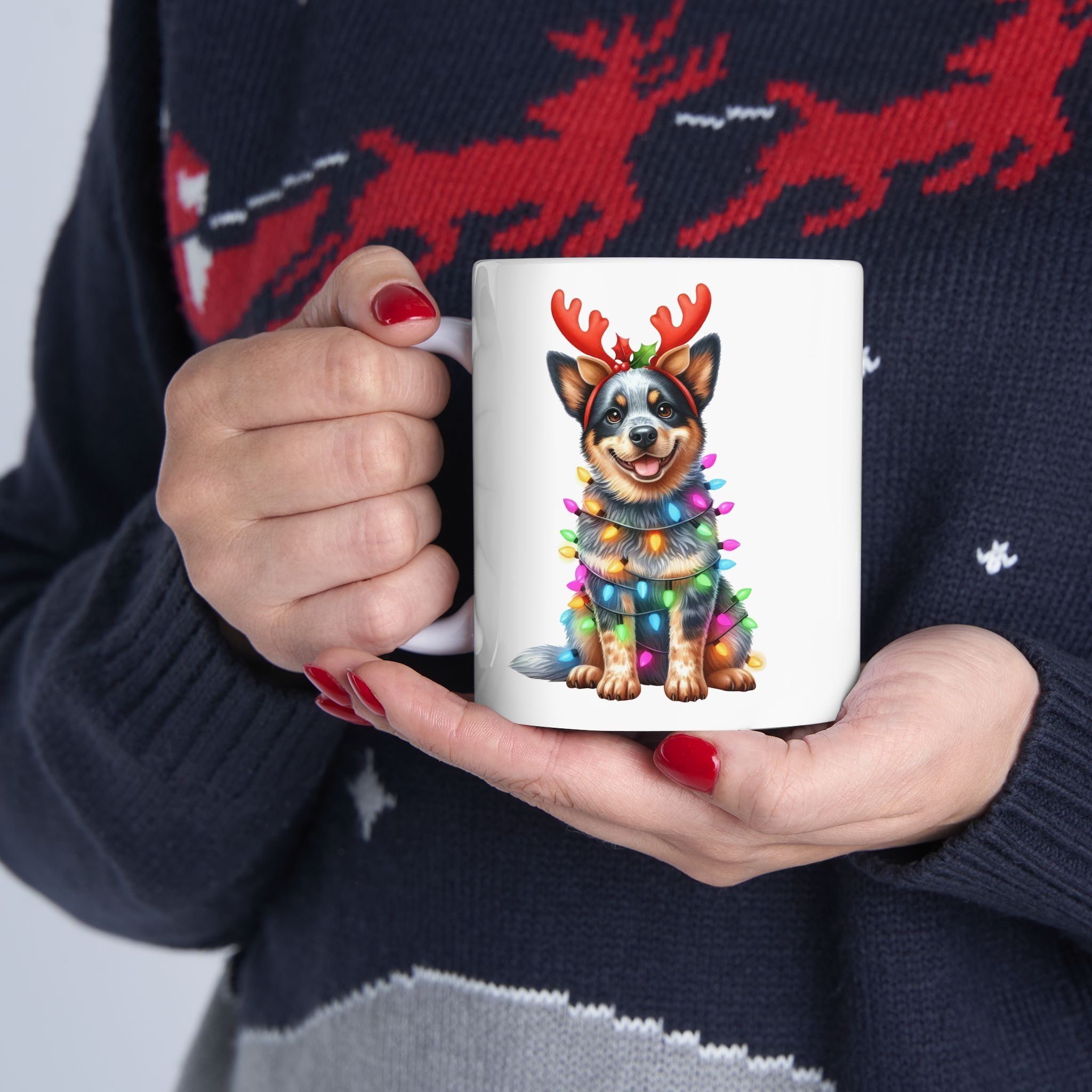 A person holds a white mug featuring an Australian Cattle Dog wearing reindeer antlers and Christmas lights.