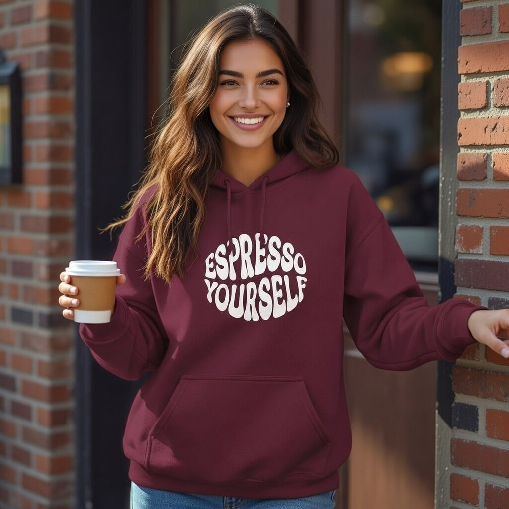 A woman smiles in a maroon Expresso Yourself hoodie while holding a coffee cup against a brick wall.