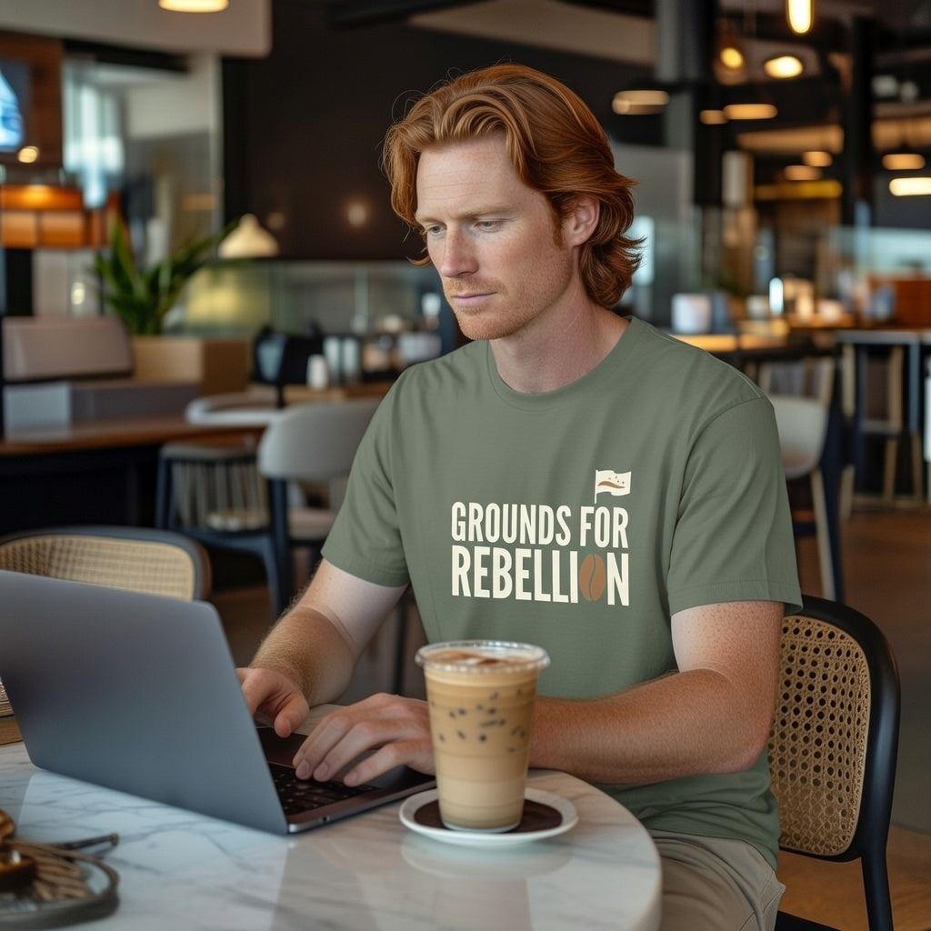 A man in a cafe wears an olive green Grounds For Rebellion t-shirt while working on a laptop.