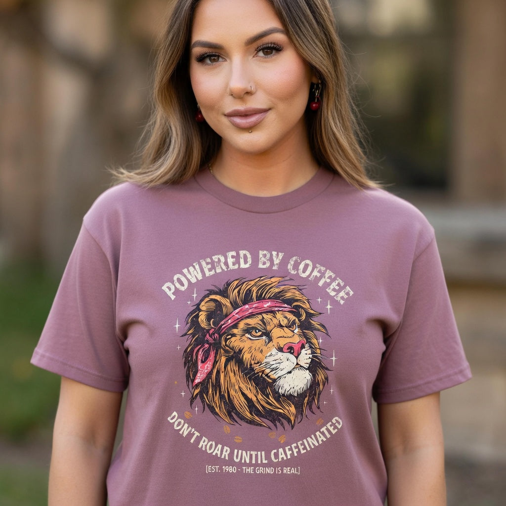 A woman wearing a mauve t-shirt with a distressed lion graphic and Powered by Coffee text.