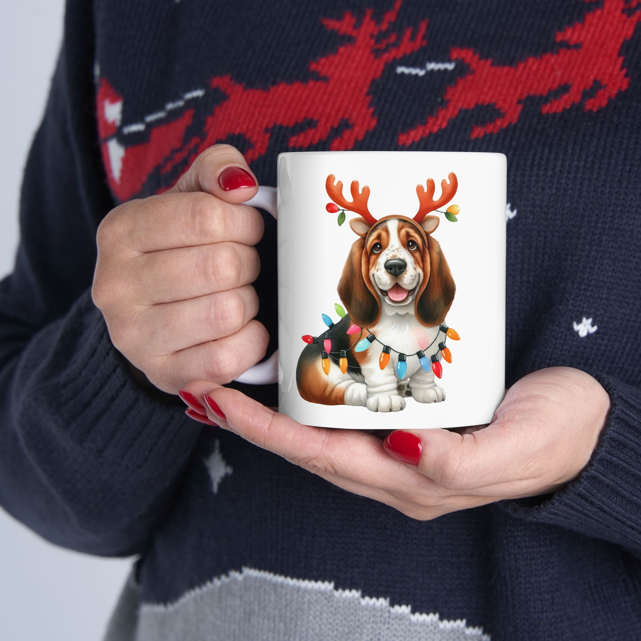 A person in a festive sweater holds a white mug with a basset hound wearing reindeer antlers.
