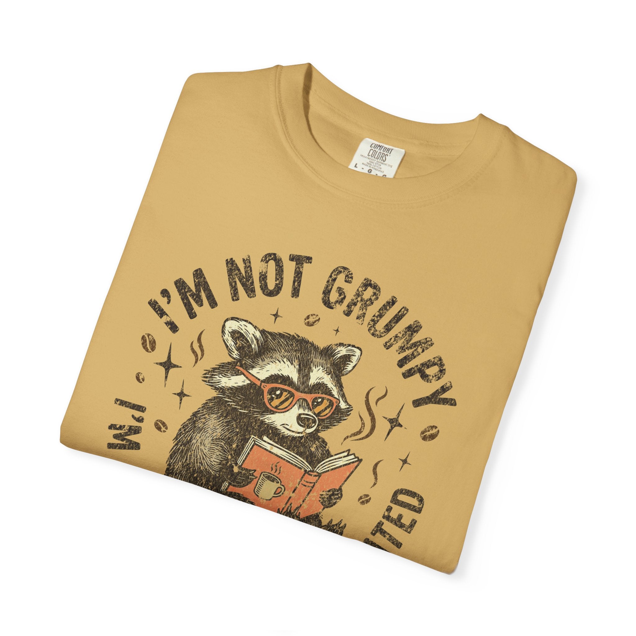 Tan Comfort Colors t-shirt featuring a raccoon wearing glasses and reading a book with coffee.