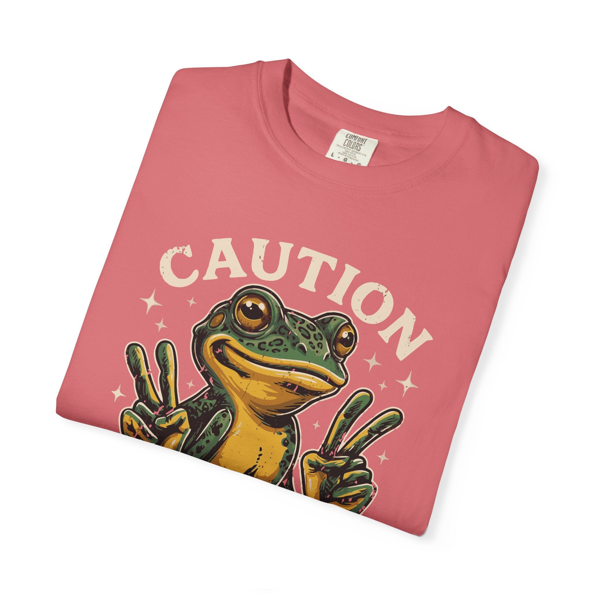 A folded pink t-shirt featuring a graphic of a smiling frog making peace signs with both hands.