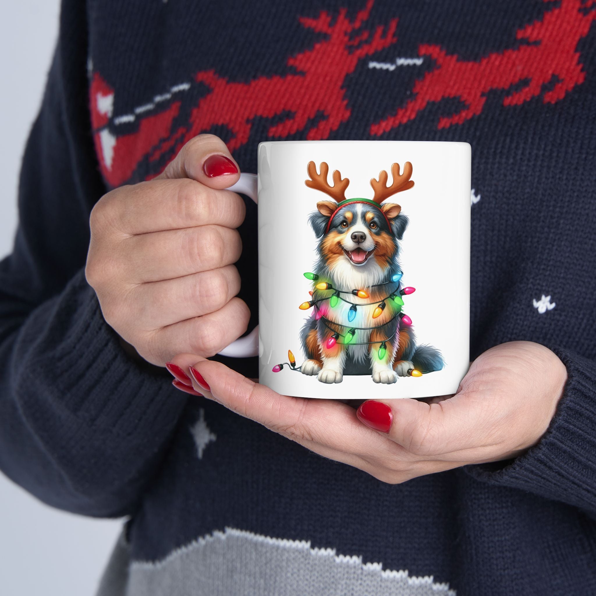 A person in a festive sweater holds a white mug with a smiling Australian Shepherd wearing antlers and lights.