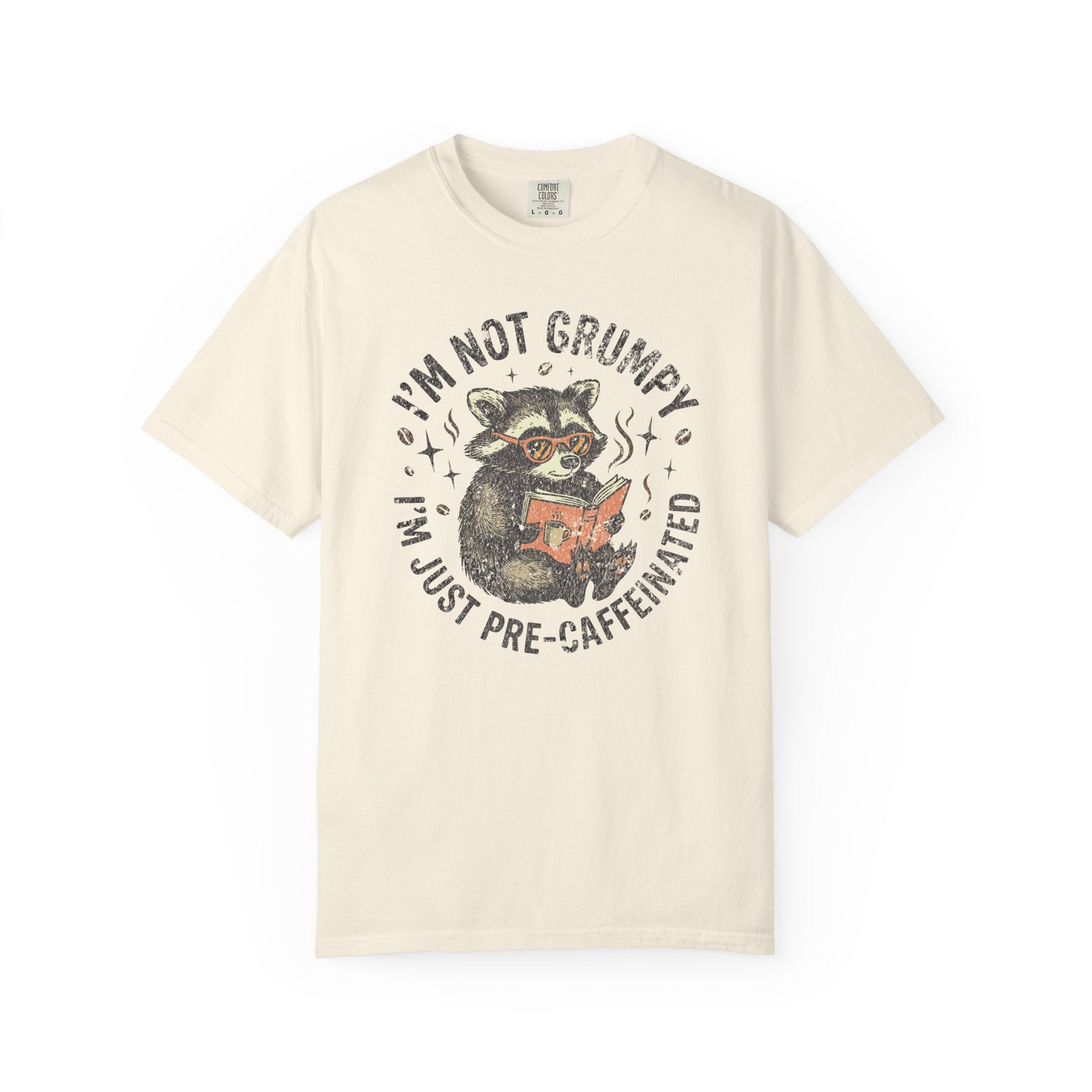 Cream-colored unisex t-shirt featuring a graphic of a raccoon reading a book with coffee.