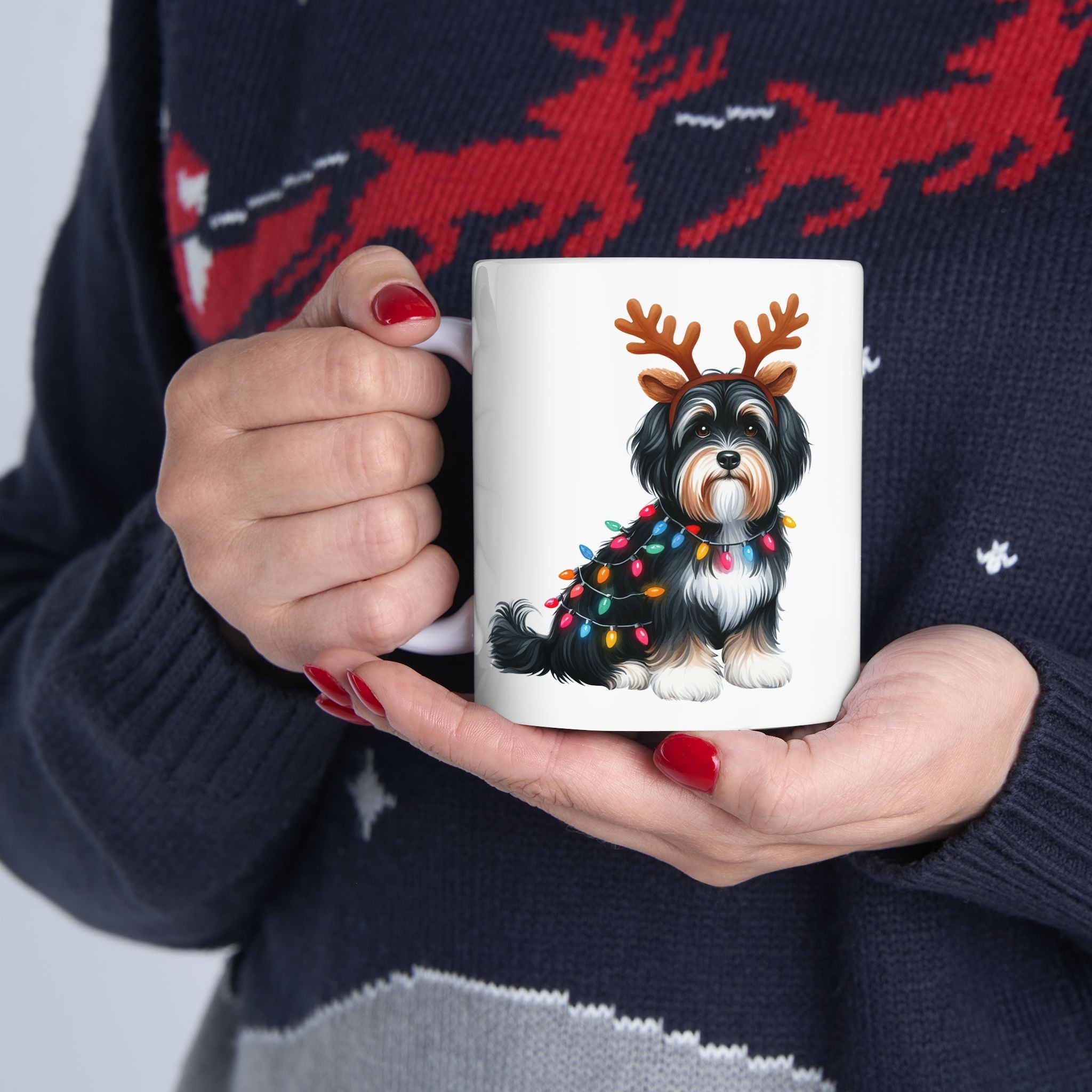 A person in a festive sweater holds a white mug with a Havanese dog wearing reindeer antlers.