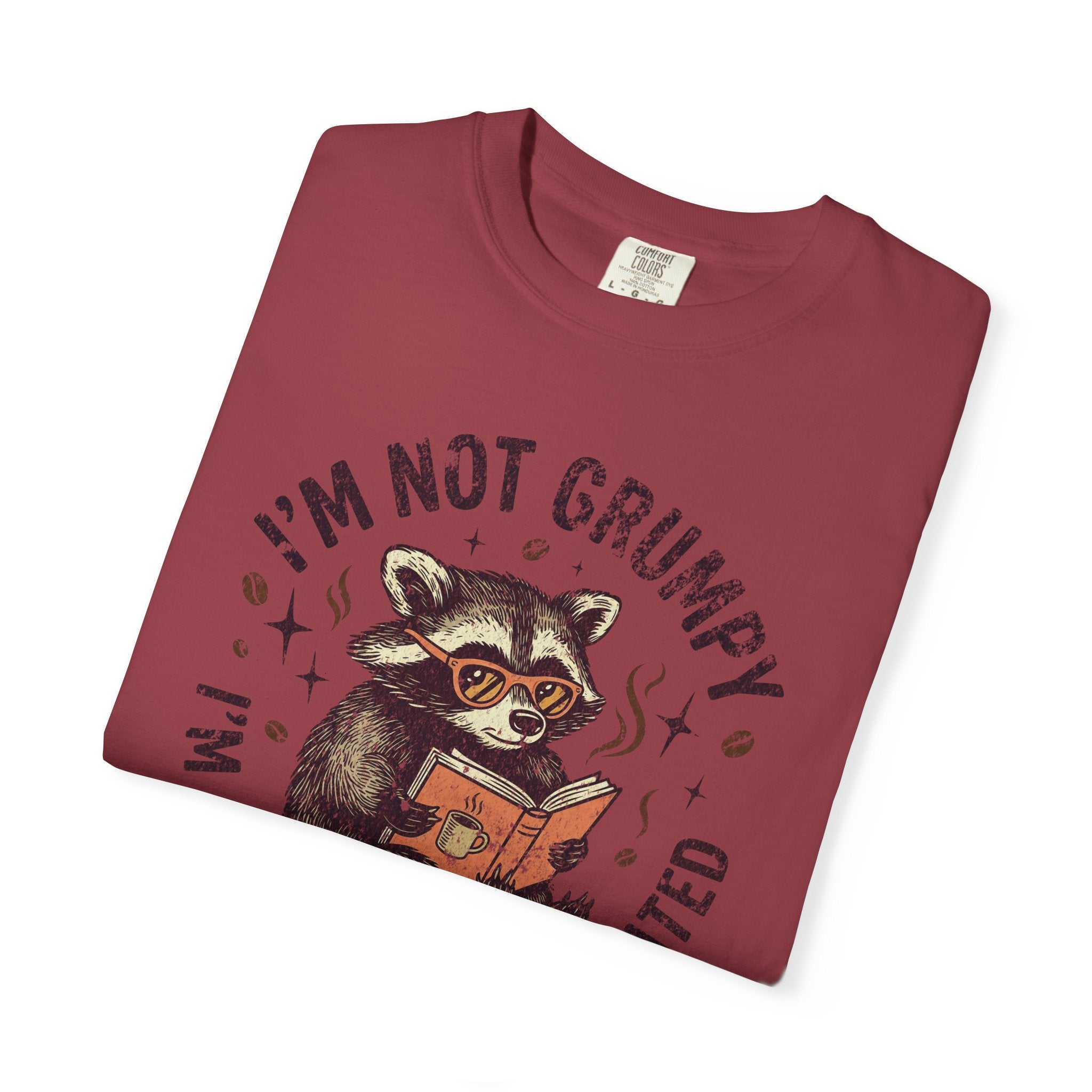A folded maroon t-shirt featuring a graphic of a raccoon wearing glasses and reading a book.
