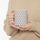 A person in a beige sweater holds a white ceramic mug featuring a repeating heart-steam pattern.