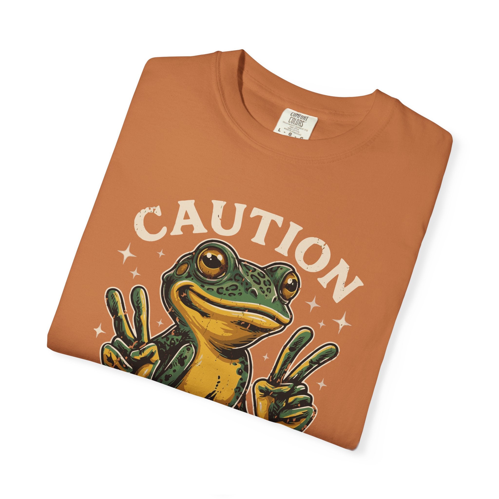 Folded orange t-shirt featuring a green frog making peace signs with the text CAUTION above it.