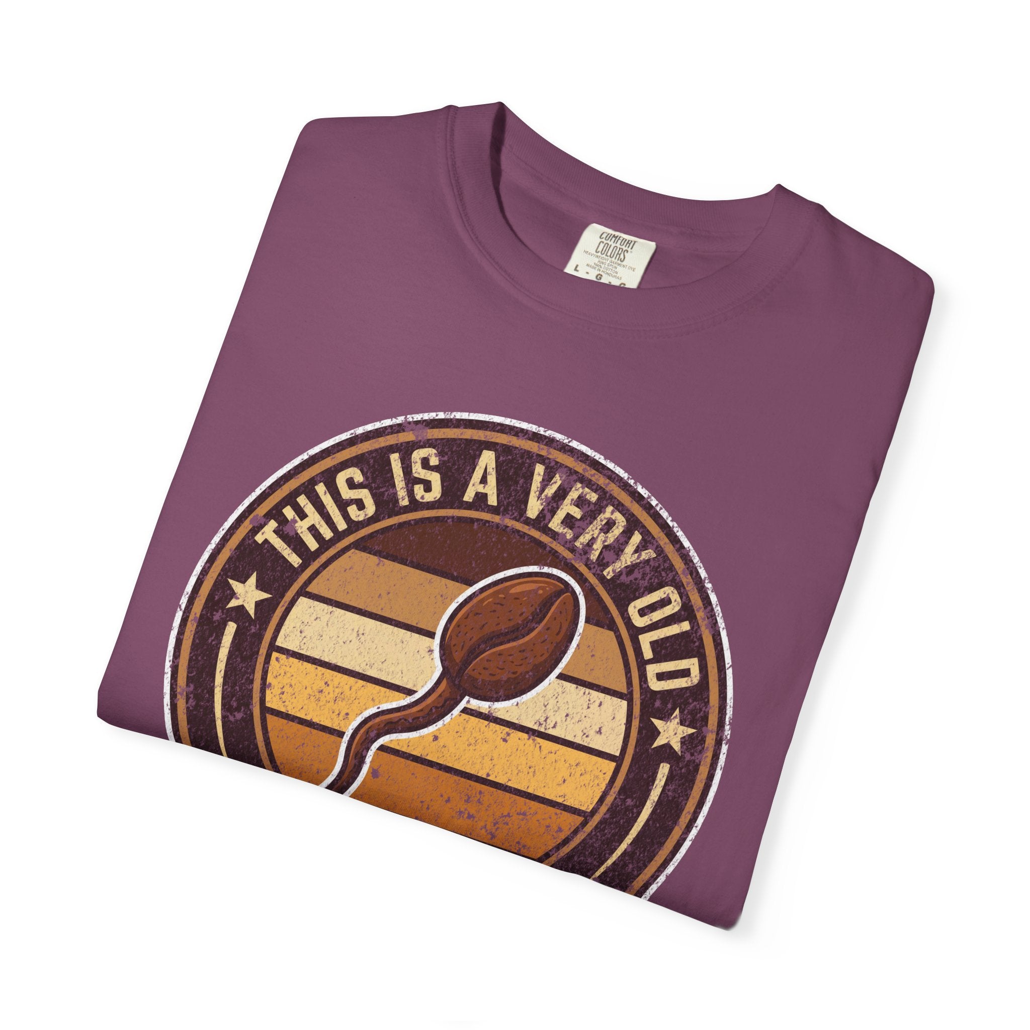 A folded purple t-shirt with a vintage-style graphic of a coffee bean and the text This Is A Very Old