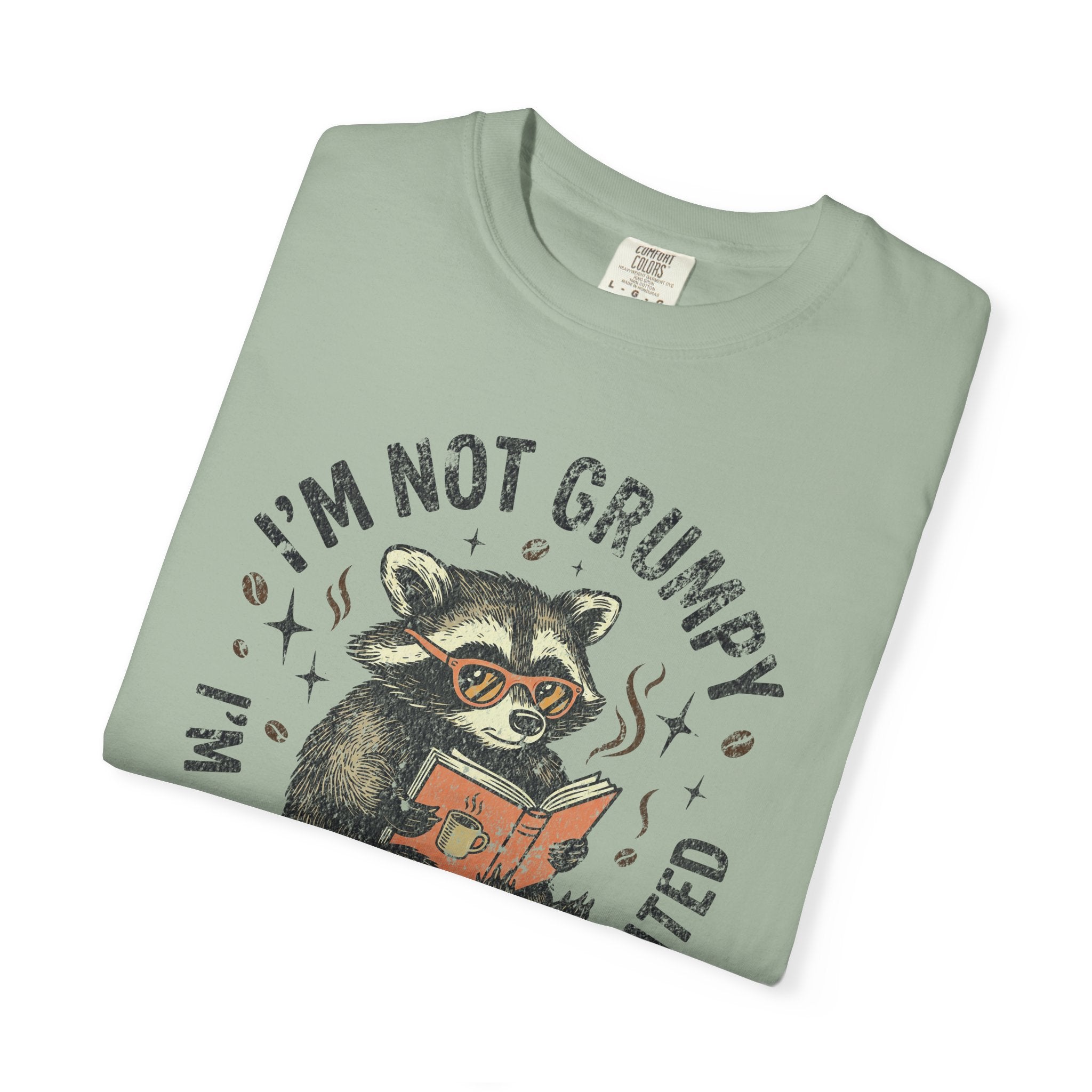 Light green unisex t-shirt featuring a graphic of a raccoon reading a book with coffee.