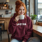 A woman wearing a maroon hoodie with retro bubble letters that say Coffee while holding a mug.