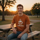 A man wearing a burnt orange Coffee Time t-shirt with a cute steaming cup graphic in a park.