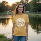 A woman wearing a mustard yellow Espresso Yourself t-shirt stands outdoors by a lake at sunset.