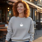 A woman smiles in a light gray pullover featuring a minimalist white steaming coffee cup graphic.