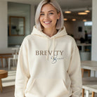 Minimalist cream-colored Brevity Brew hoodie with a script logo worn by a smiling woman.