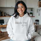 A woman smiles in a white Brevity Brew minimalist script logo hoodie with a front pocket.
