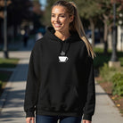 A woman smiles in a black hoodie featuring a small white steaming coffee cup graphic on the chest.