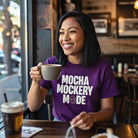 A woman in a cafe wears a purple Mocha Mockery Mode t-shirt while holding a cup of coffee.