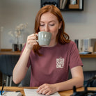 A woman in a mauve Brewed Chaos Only t-shirt drinks from a mug while sitting at a desk.