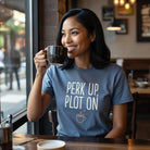 A woman in a blue Perk Up, Plot On t-shirt smiles while holding a coffee mug in a cafe.