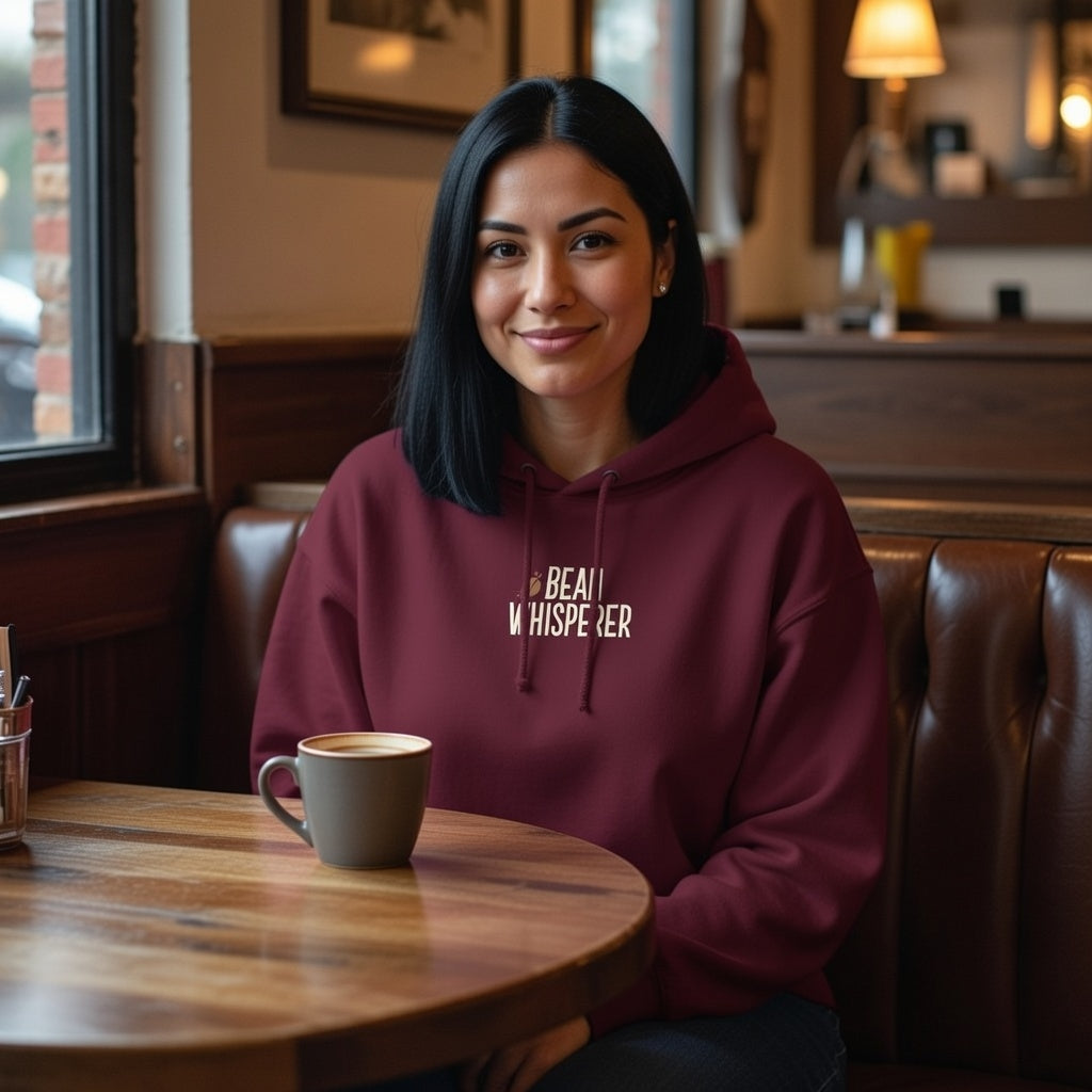 A woman in a cafe wearing a maroon Bean Whisperer hoodie while sitting at a table with coffee.