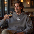 A man in a cafe wears a grey Bean Whisperer hoodie while holding a small glass of espresso.