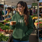A woman wears a dark green Filterless Fuel hoodie while holding a coffee cup at an outdoor market.