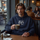 A man in a cafe wearing a navy blue Sip Of Sanity hoodie while holding a cup of coffee.