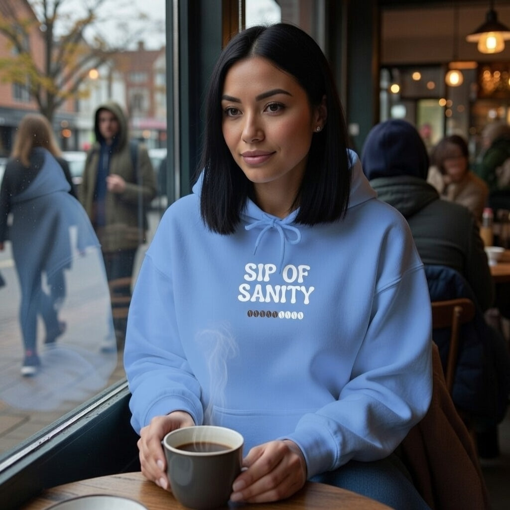 A woman in a light blue Sip Of Sanity hoodie sits in a cafe holding a steaming cup of coffee.