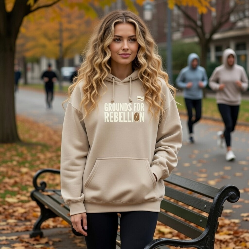 A woman wears a beige Grounds For Rebellion hoodie with a coffee bean graphic in an autumn park.