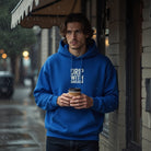 A man in a blue hoodie with Drip With Sarcasm printed on the front holds a coffee cup in the rain.