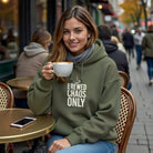 A woman wears an olive green Brewed Chaos Only hoodie while holding a cup of coffee at a cafe.