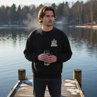 A man on a dock wearing a black Latte Night Mayhem sweatshirt while holding a steaming travel mug.