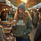 A woman wears a green Bean Whisperer sweatshirt while holding a coffee cup at an outdoor market.