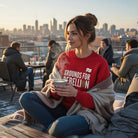 A woman wears a red Grounds For Rebellion coffee sweatshirt while holding a steaming mug outdoors.