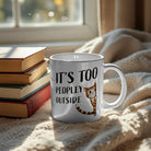 White ceramic mug with a peeking cat and the text It's Too Peopley Outside next to a stack of books.