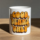 White ceramic mug with retro 70s style Good Beans Only text and floral accents on a wooden surface.