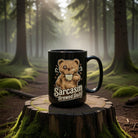 Black mug with a teddy bear and Sarcasm Brewed Daily text on a mossy tree stump in a forest.