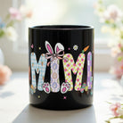 Black ceramic mug featuring the word MIMI in colorful pastel Easter patterns with bunny ears and bow.