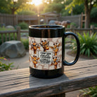 Black ceramic coffee mug featuring cartoon giraffes and a sarcastic quote on a wooden railing.