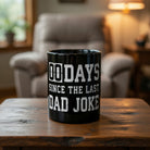 Black coffee mug with white text reading 00 Days Since The Last Dad Joke on a wooden table.