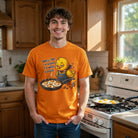 A man in a kitchen wears an orange t-shirt with a sleepy chick graphic and Brew-Tal! coffee pun.