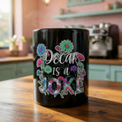 Black ceramic mug with floral designs and the text Decaf is a Joke on a wooden table.