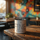 White ceramic mug with 'I'm Fine... But Need Coffee' word cloud design on a rustic wooden table.